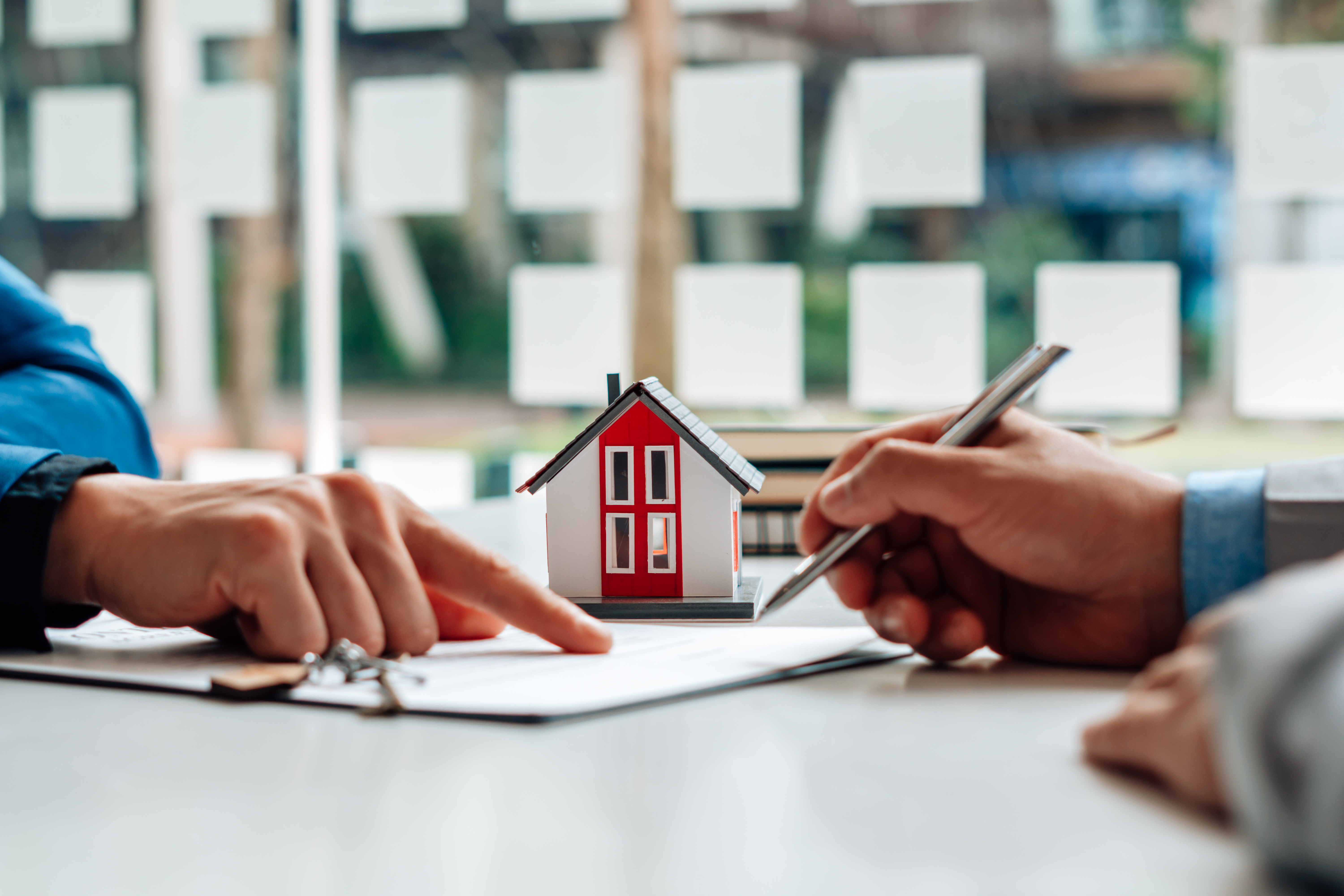 Two hands over a document, one holding a pen, with a toy house in the background.
