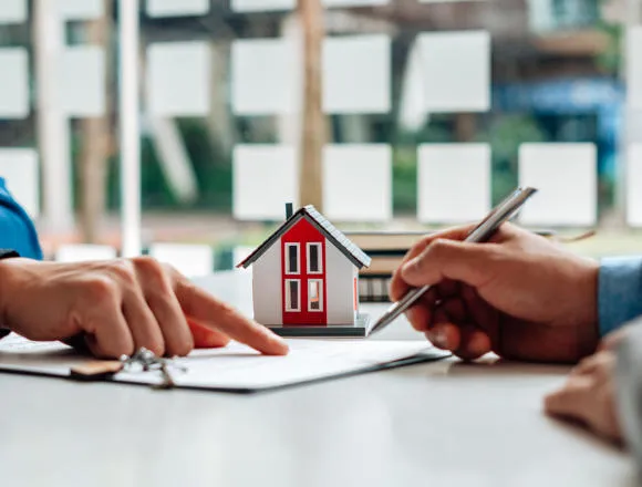 Two hands over a document, one holding a pen, with a toy house in the background.